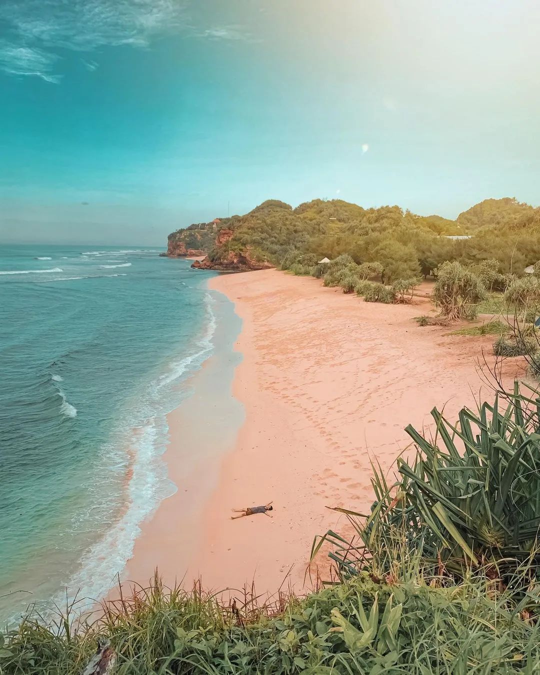 A person lying on the sandy shore of Watu Kodok Beach with greenery in the background.