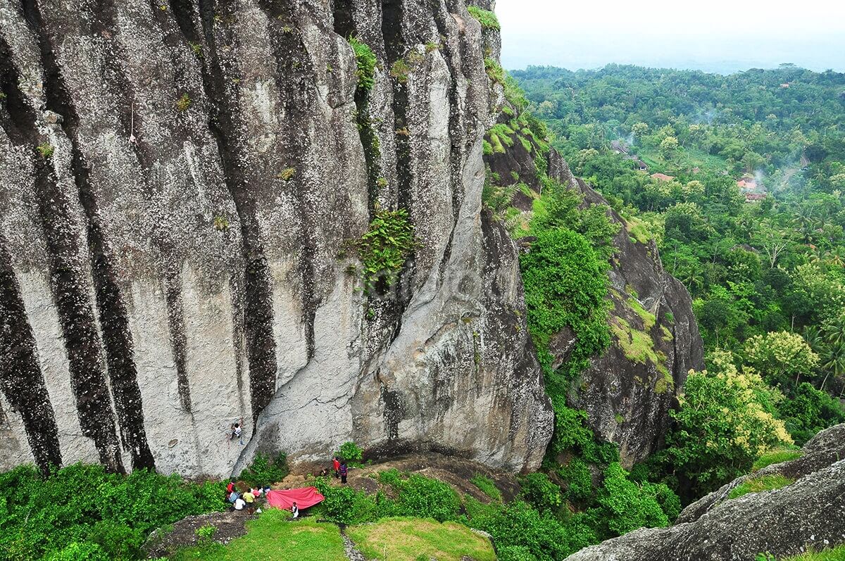 A group of climbers at the base of a towering rock face on Nglanggeran Mountain with lush green surroundings.