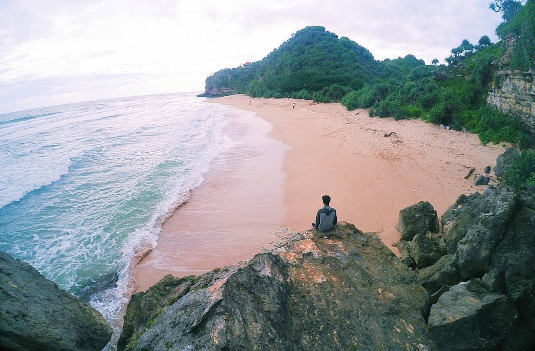 A person sitting on a rock, overlooking the quiet and scenic Sanglen Beach with its smooth sand and green hills.