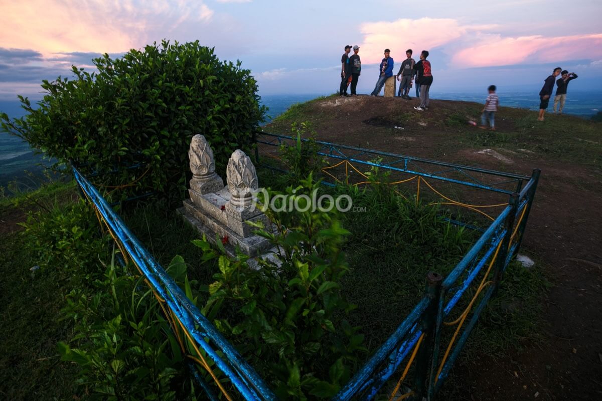 A small shrine with stone carvings surrounded by grass, with people gathering nearby on a hilltop during sunset.