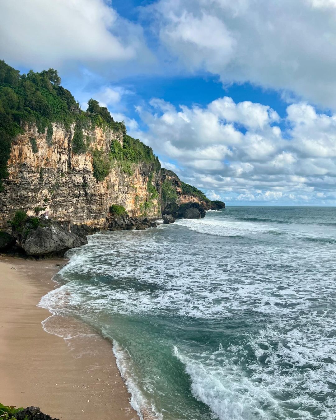 A view of Ngrumput Beach with waves crashing against the shore, backed by tall green cliffs under a blue sky with white clouds.