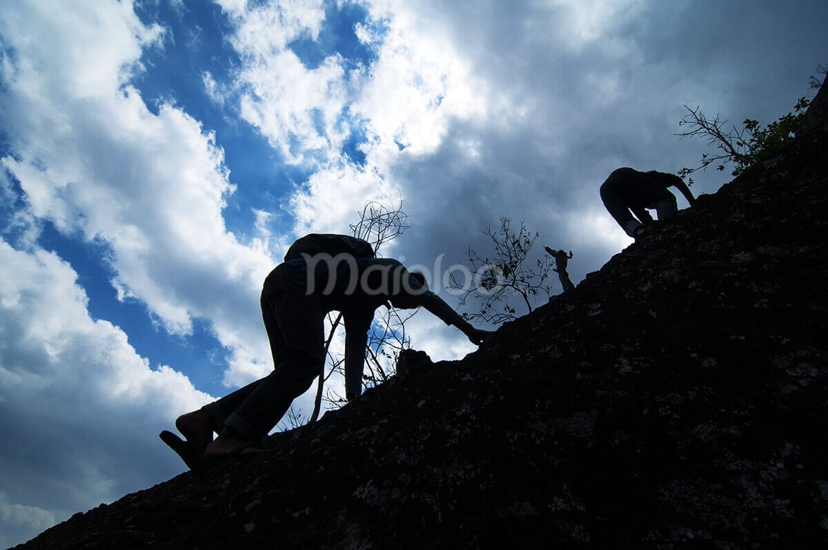 Two climbers scrambling up a steep rocky slope with a dramatic sky in the background on Nglanggeran Mountain.