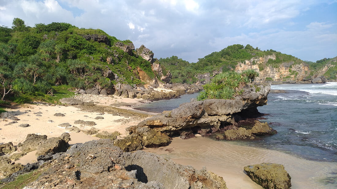 A secluded beach with rocky formations and green hills under a partly cloudy sky.