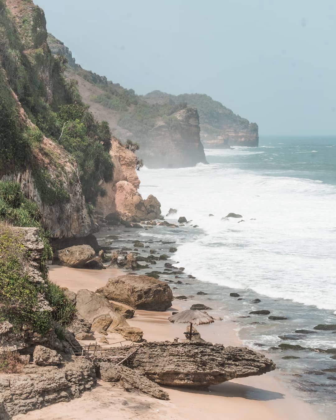 The coastline of Seruni Beach with cliffs, rocky formations, and waves crashing onto the sandy shore.