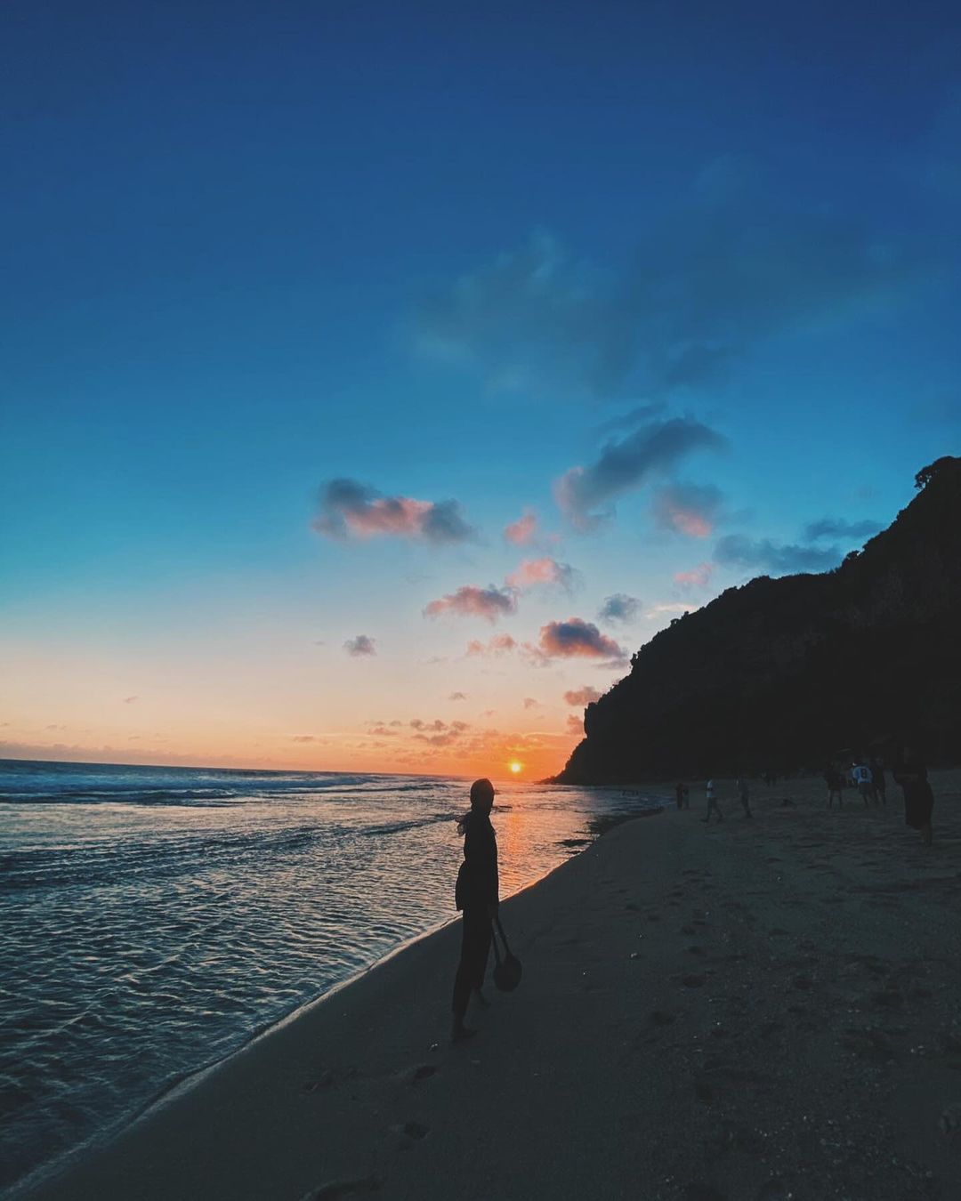 A person standing on the shoreline at sunset at Watunene Beach, with the sun setting behind the karst cliffs.