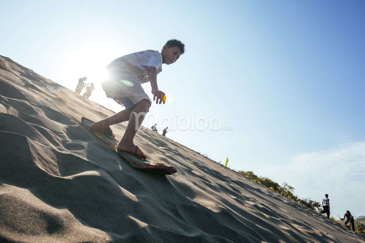 A young boy sandboarding down a dune with the sun shining brightly behind him.