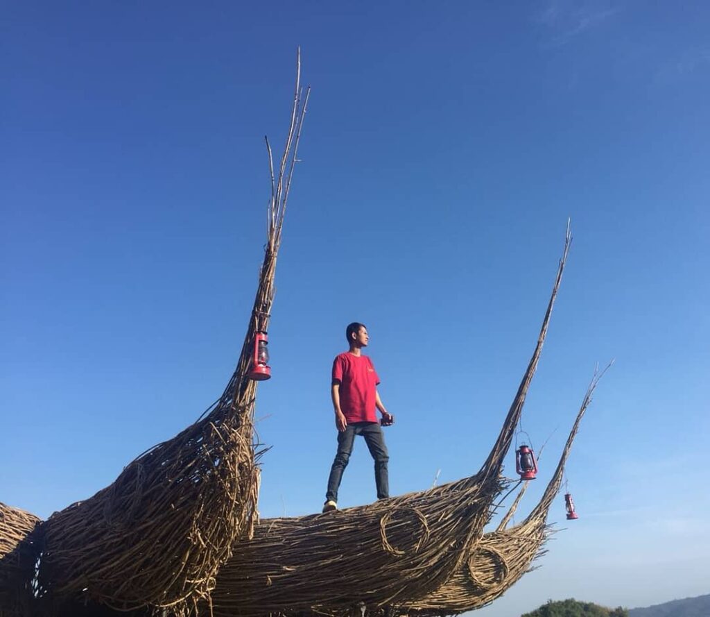 A person stands confidently on top of a wicker art installation with lanterns, under a clear blue sky at Geoforest Watu Payung Turunan.