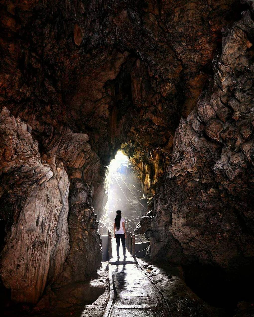 A person stands on a narrow path inside Kiskendo Cave, illuminated by sunlight streaming in from the entrance, revealing the rocky interior walls.