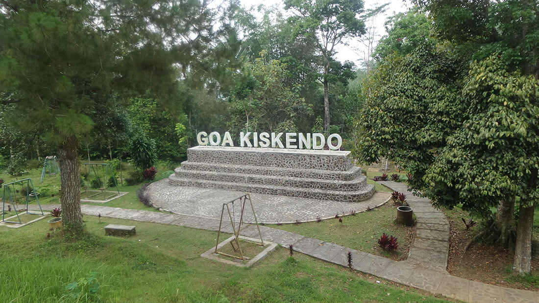 A tiered stone platform with the large sign "Goa Kiskendo" at the entrance, surrounded by greenery and trees.