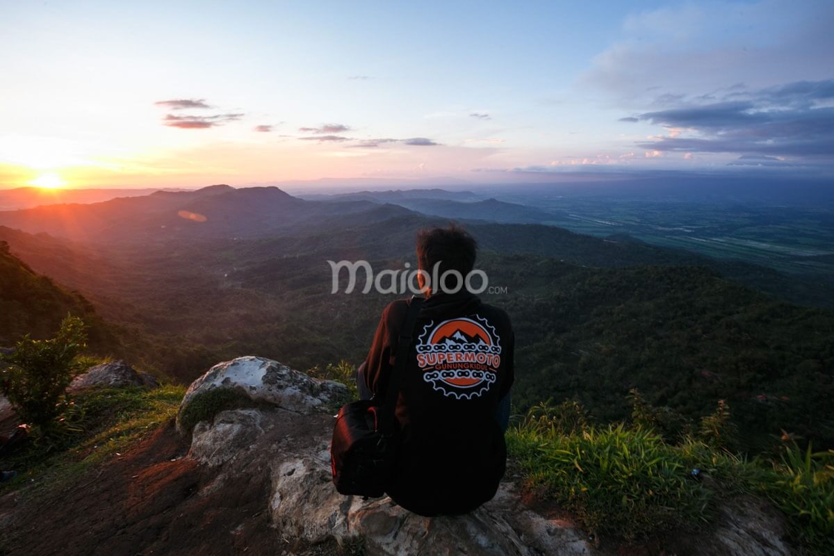 A person sitting on a hilltop, looking at a sunset over a valley with mountains in the background.