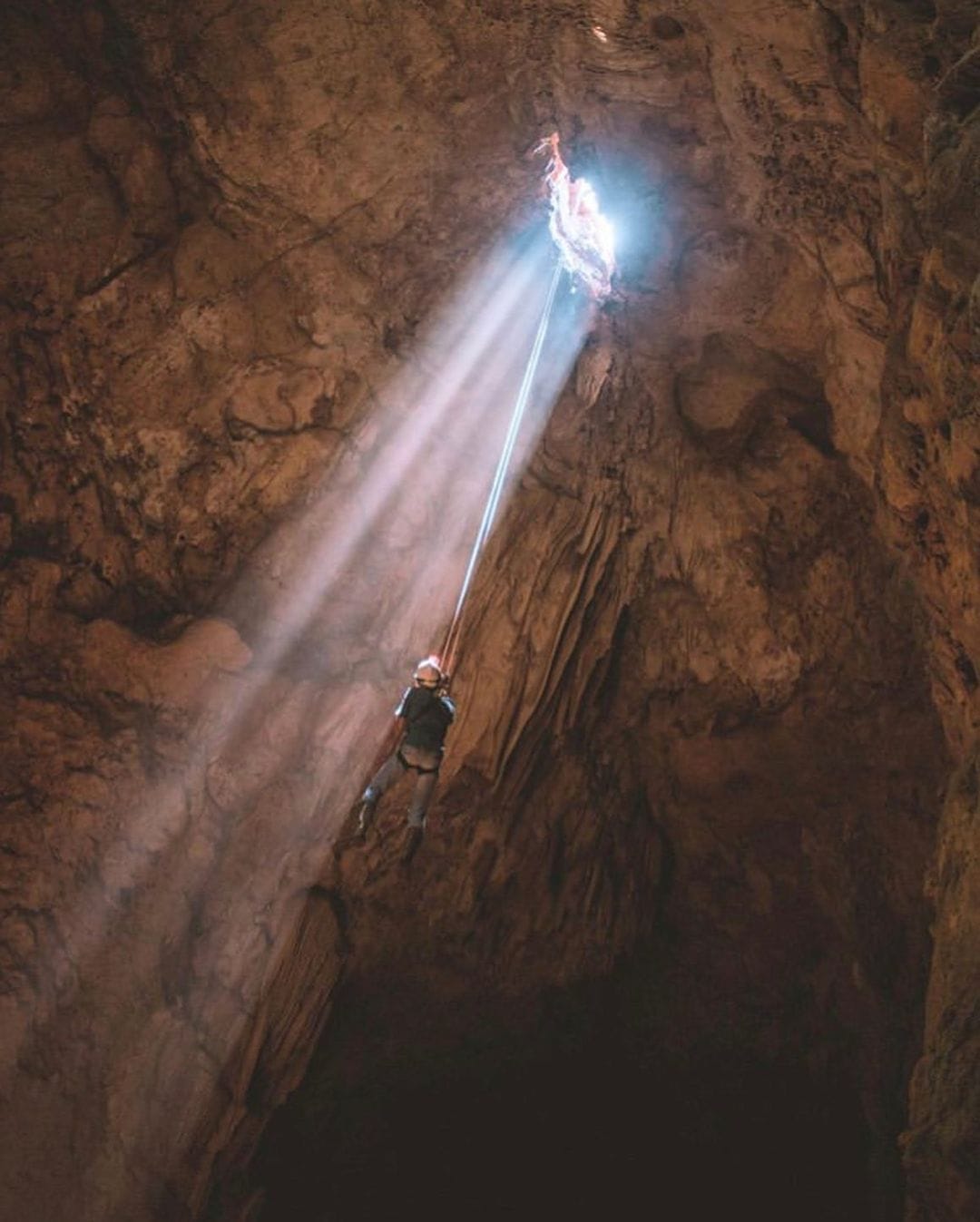 A person wearing a helmet rappels into Cokro Cave with a beam of sunlight shining through the cave's entrance above.