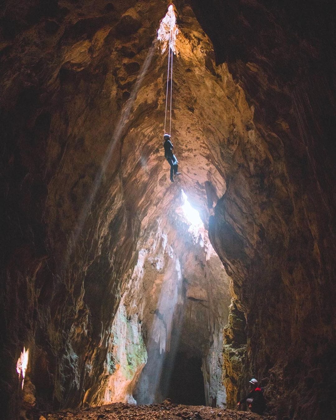 A person wearing a helmet rappels down the vertical entrance of Cokro Cave, with another person watching from below. Sunlight streams into the cave through the opening above.