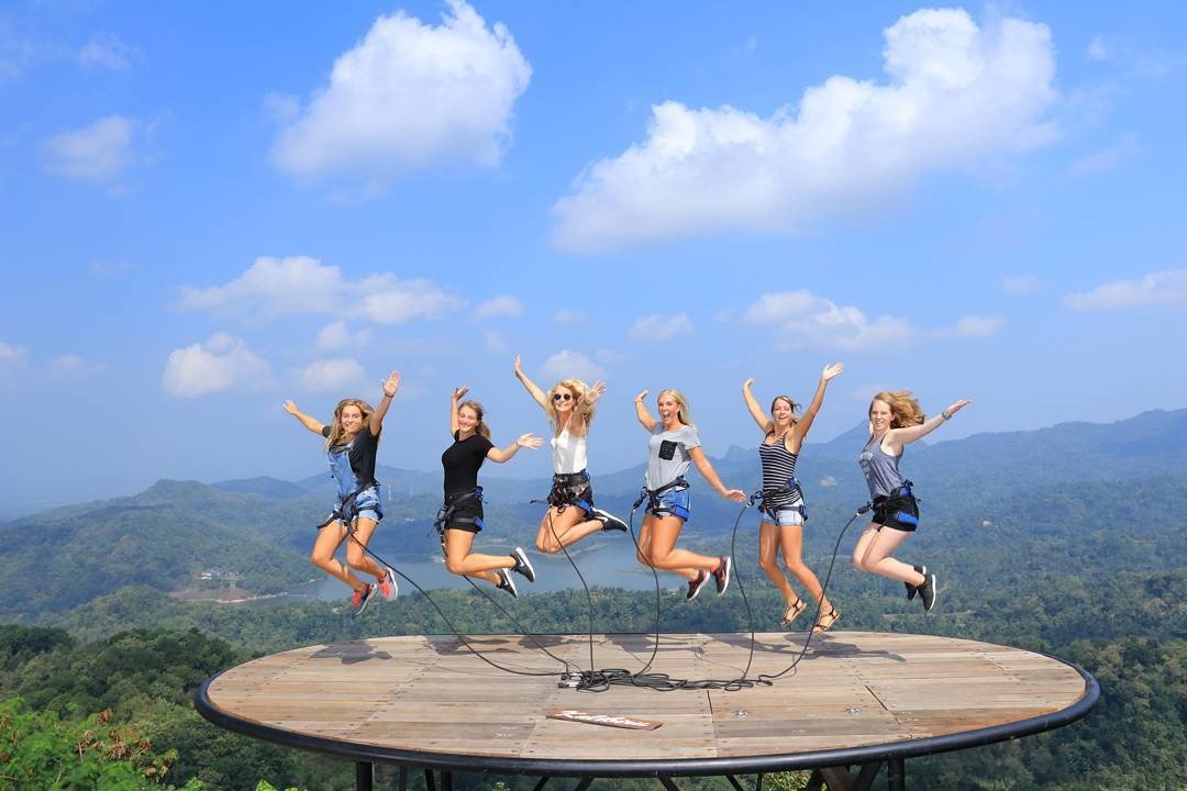 A group of people wearing harnesses, jumping joyfully on a large wooden platform with scenic views of the lush hills and Sermo Reservoir at Kalibiru Natural Tourism in Yogyakarta, Indonesia.