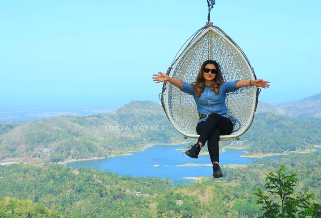 A person enjoying a relaxing ride on a large hanging swing chair, with arms outstretched and a scenic view of the lush green hills and Sermo Reservoir in the background at Kalibiru Natural Tourism in Yogyakarta, Indonesia.