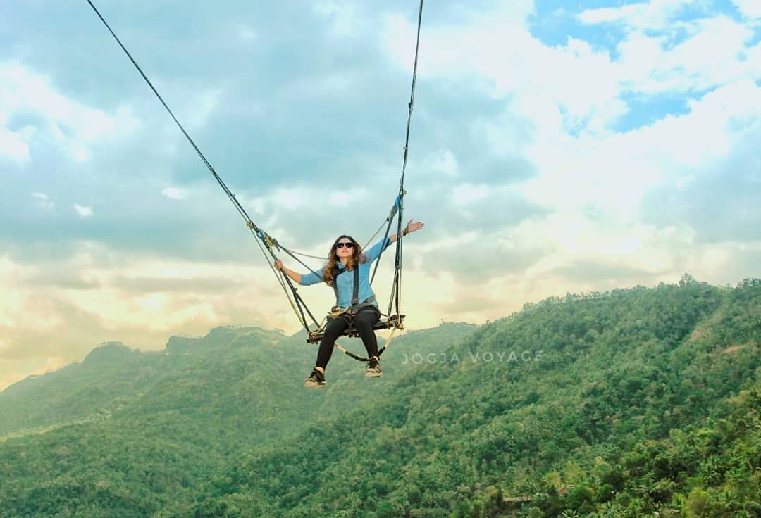 A person riding a high swing with arms outstretched, enjoying the stunning view of the lush green hills at Kalibiru Natural Tourism in Yogyakarta, Indonesia.