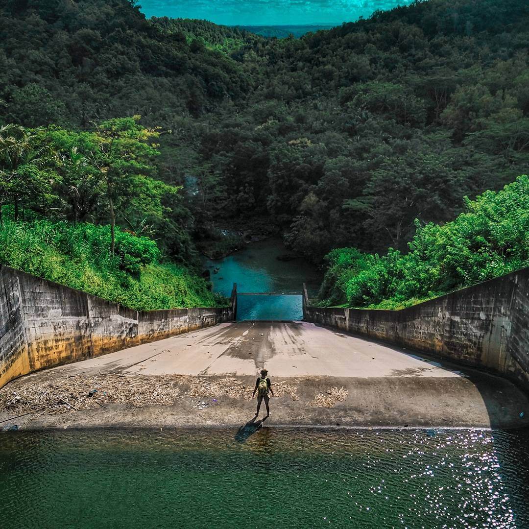 A person stands at the end of a dam outlet at Sermo Reservoir, surrounded by dense green forest.