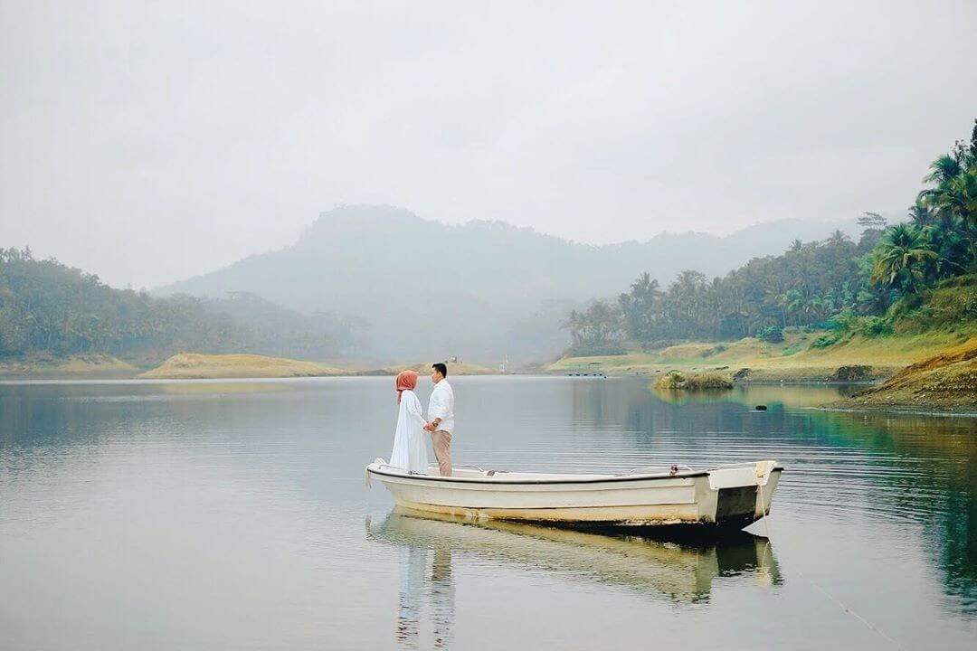 A couple stands on a small boat in the calm waters of Sermo Reservoir with lush green hills in the background.