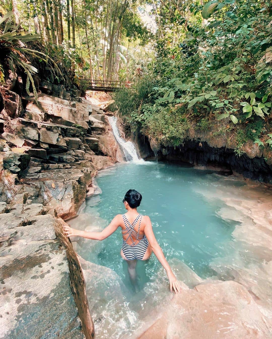 A person in a striped swimsuit stepping into a turquoise pool at Kedung Pedut Waterfall, surrounded by rock formations and lush greenery.