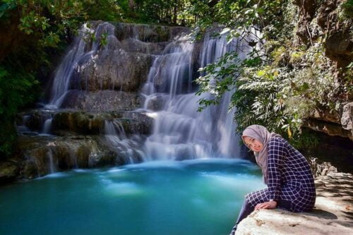 A woman sitting by the turquoise pool of Lepo Waterfall in Yogyakarta, with cascading waters and lush greenery in the background.