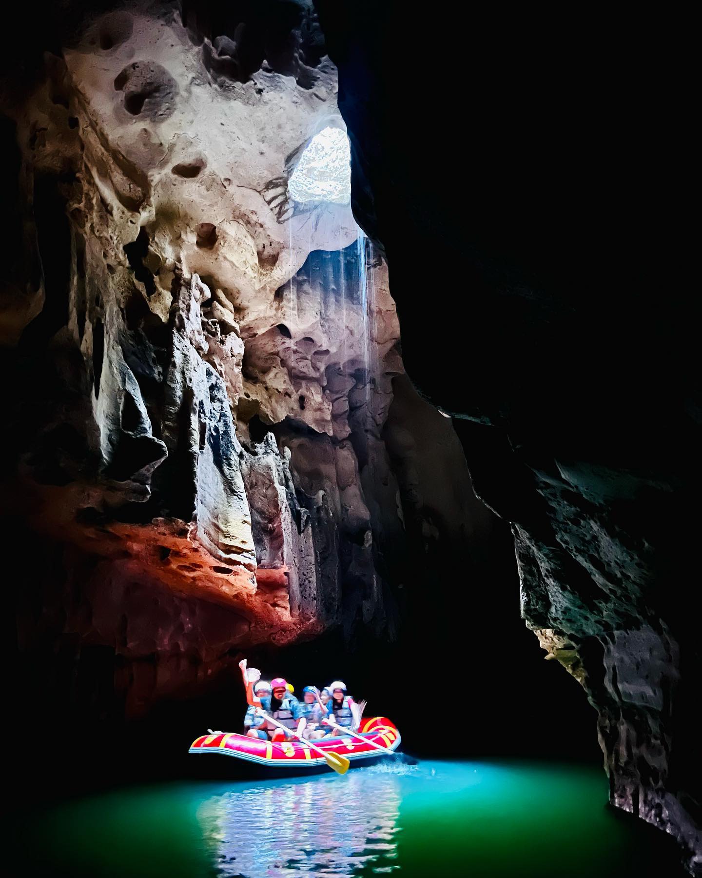 A group of people in an inflatable raft paddle through the underground river in Tanding Cave, illuminated by a beam of light shining through a hole in the cave ceiling.