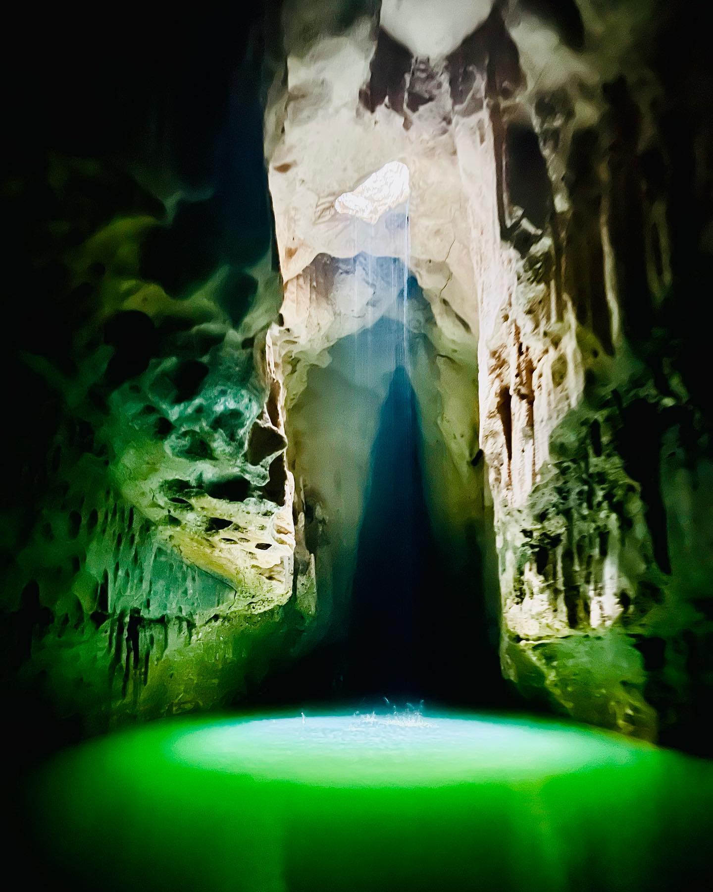 Sunlight beams through an opening in Tanding Cave’s ceiling, illuminating the green water of the underground river and casting dramatic shadows on the cave walls.