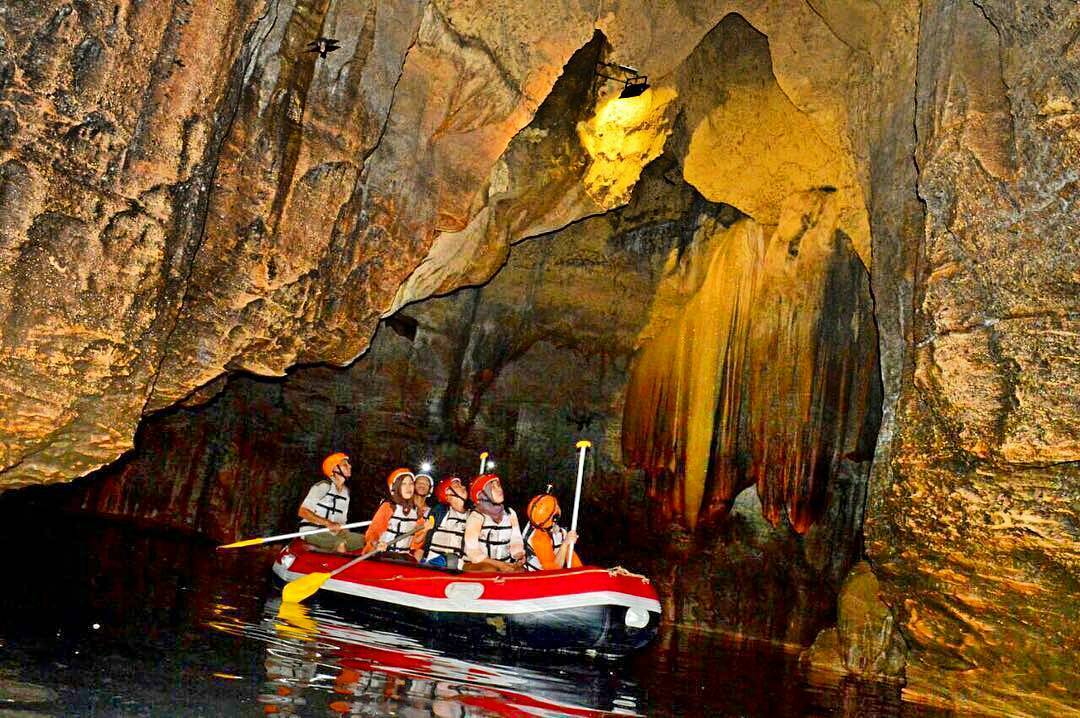 Tourists in a red inflatable raft navigate the underground river in Tanding Cave, observing the impressive stalactites and rock formations.