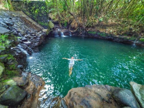 A person floating on their back in the clear blue waters of Tirta Budi Bathing Place surrounded by lush green vegetation and rocky formations.