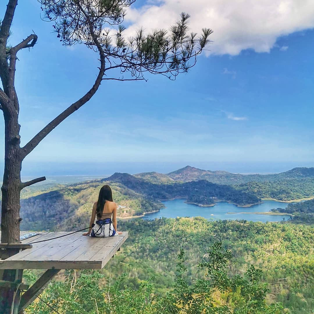 A person sitting on a wooden platform, overlooking the lush green hills and Sermo Reservoir at Kalibiru Natural Tourism in Yogyakarta, Indonesia.