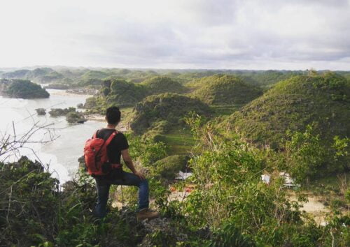 A hiker with a red backpack stands on a rock at Kosakora Peak, overlooking lush green hills and the ocean.