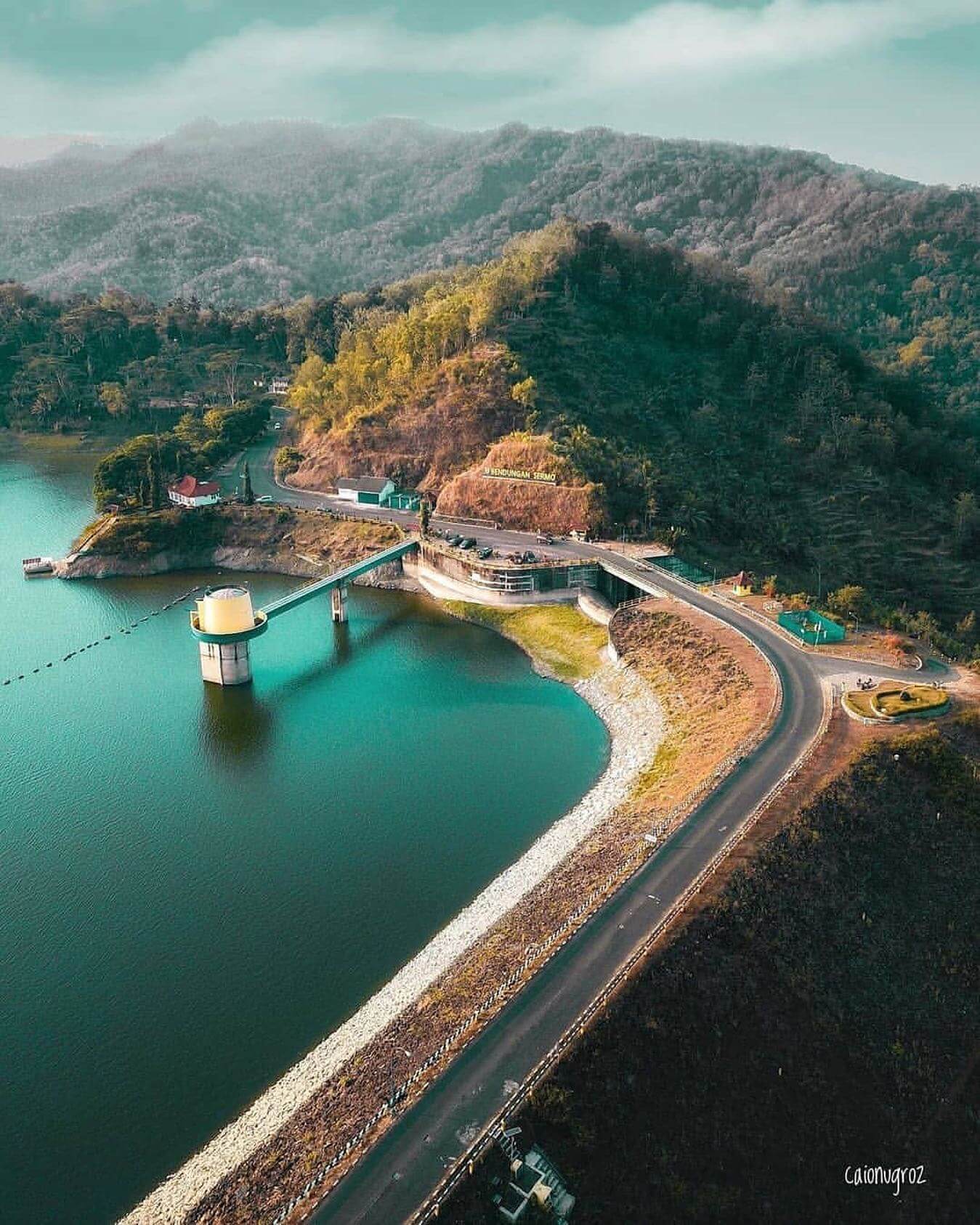 Aerial view of Sermo Reservoir surrounded by lush green mountains, featuring a road and dam structure.