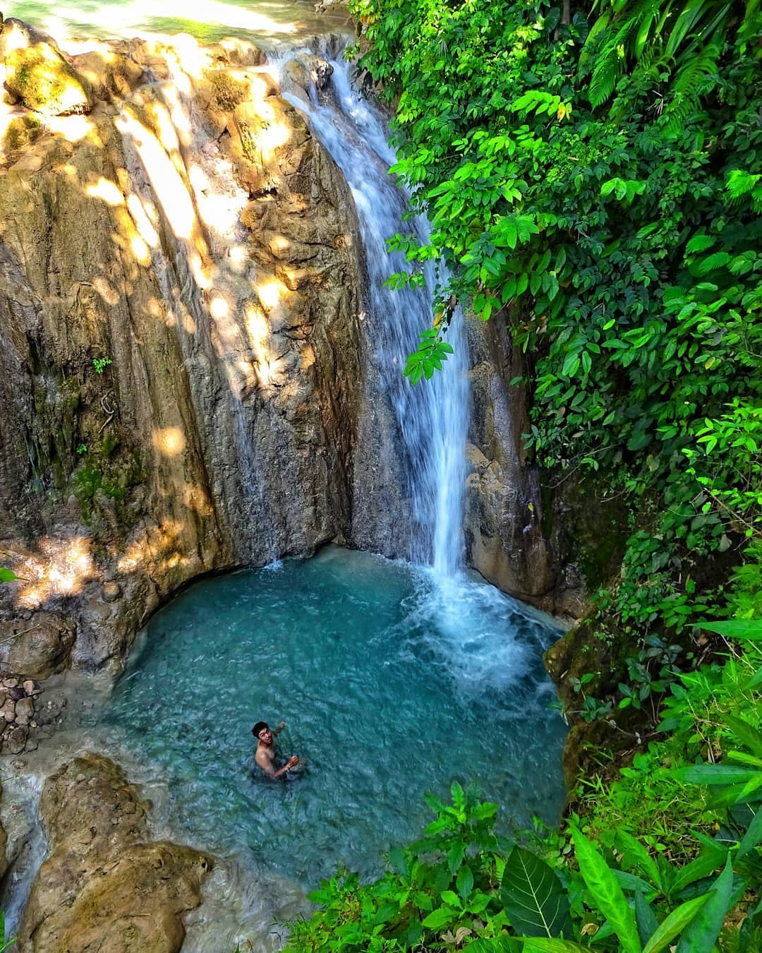 A person swimming in a turquoise pool at the bottom of Kedung Pedut Waterfall, encompassed by lush green vegetation and rocky cliffs.