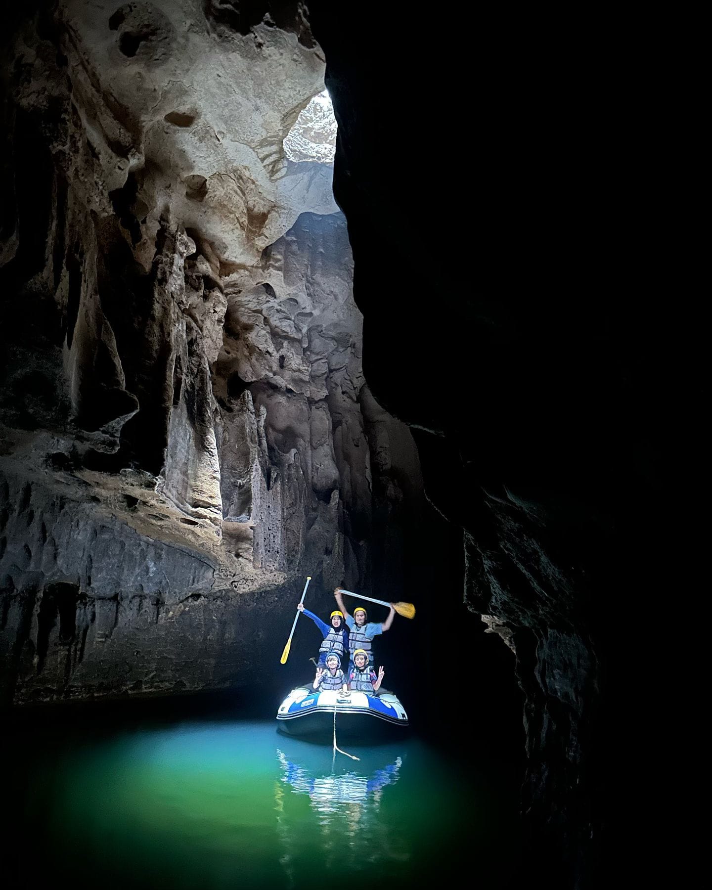 A group of people in a blue inflatable raft cheerfully paddle through the underground river in Tanding Cave, with light shining down from an opening in the cave ceiling and illuminating the green water.