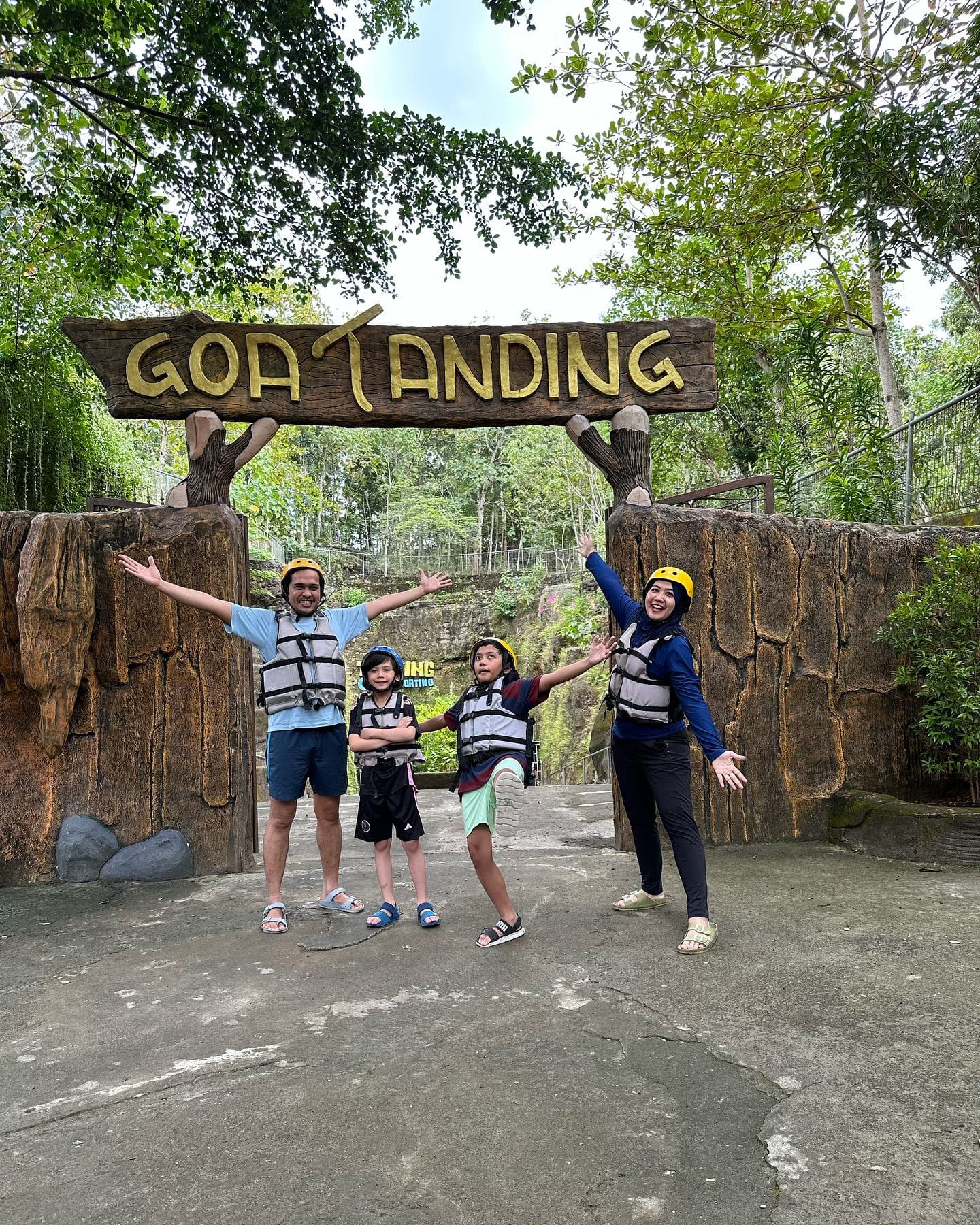 A family poses happily in front of the "Goa Tanding" sign, wearing life jackets and helmets, ready for their cave adventure.