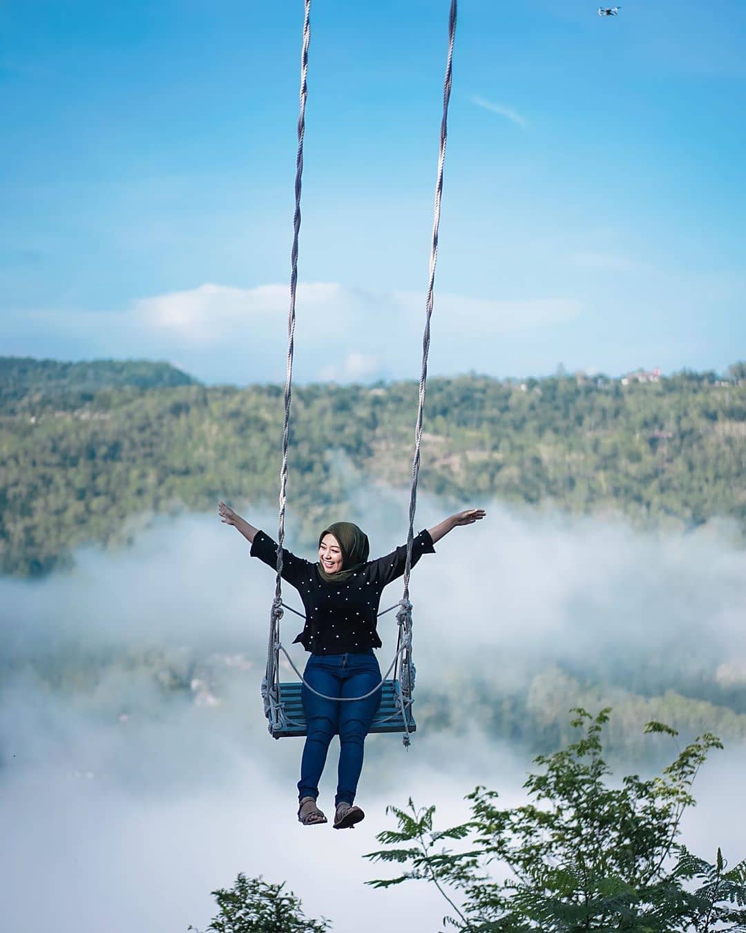 A woman joyfully swinging high above a sea of clouds with lush green hills in the background at Mojo Gumelem Hill in Yogyakarta, Indonesia.