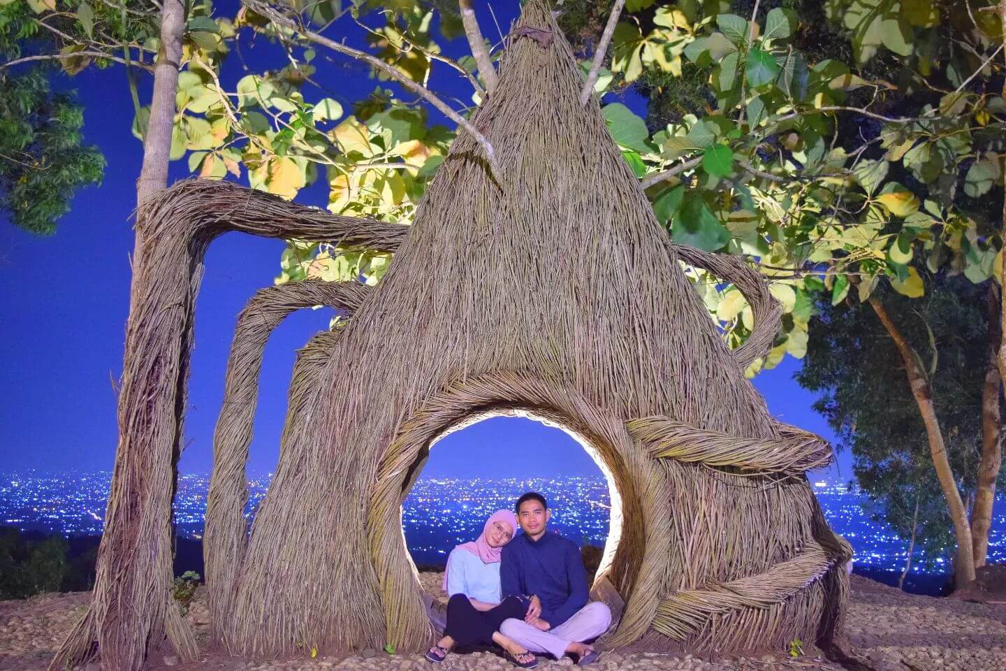 A couple sitting in a woven tree structure at Hutan Pinus Pengger with city lights in the background.