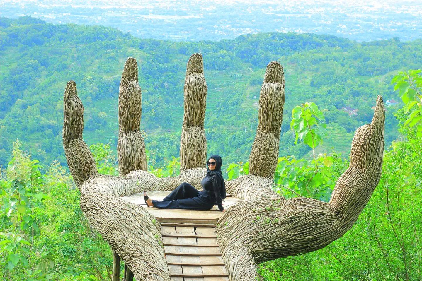 Woman relaxing on the Giant Hand installation at Hutan Pinus Pengger with lush green hills in the background.