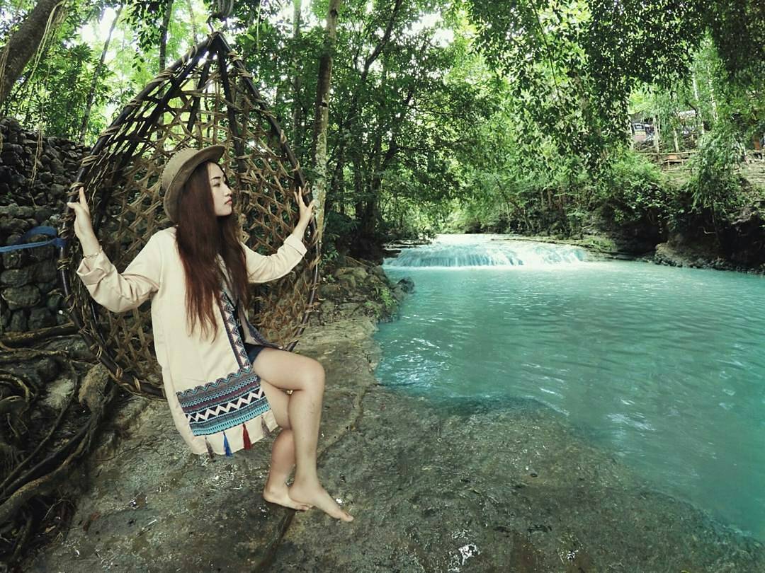 A woman sitting on a wicker swing next to the tranquil turquoise waters of Lepo Waterfall, surrounded by lush greenery in Yogyakarta.