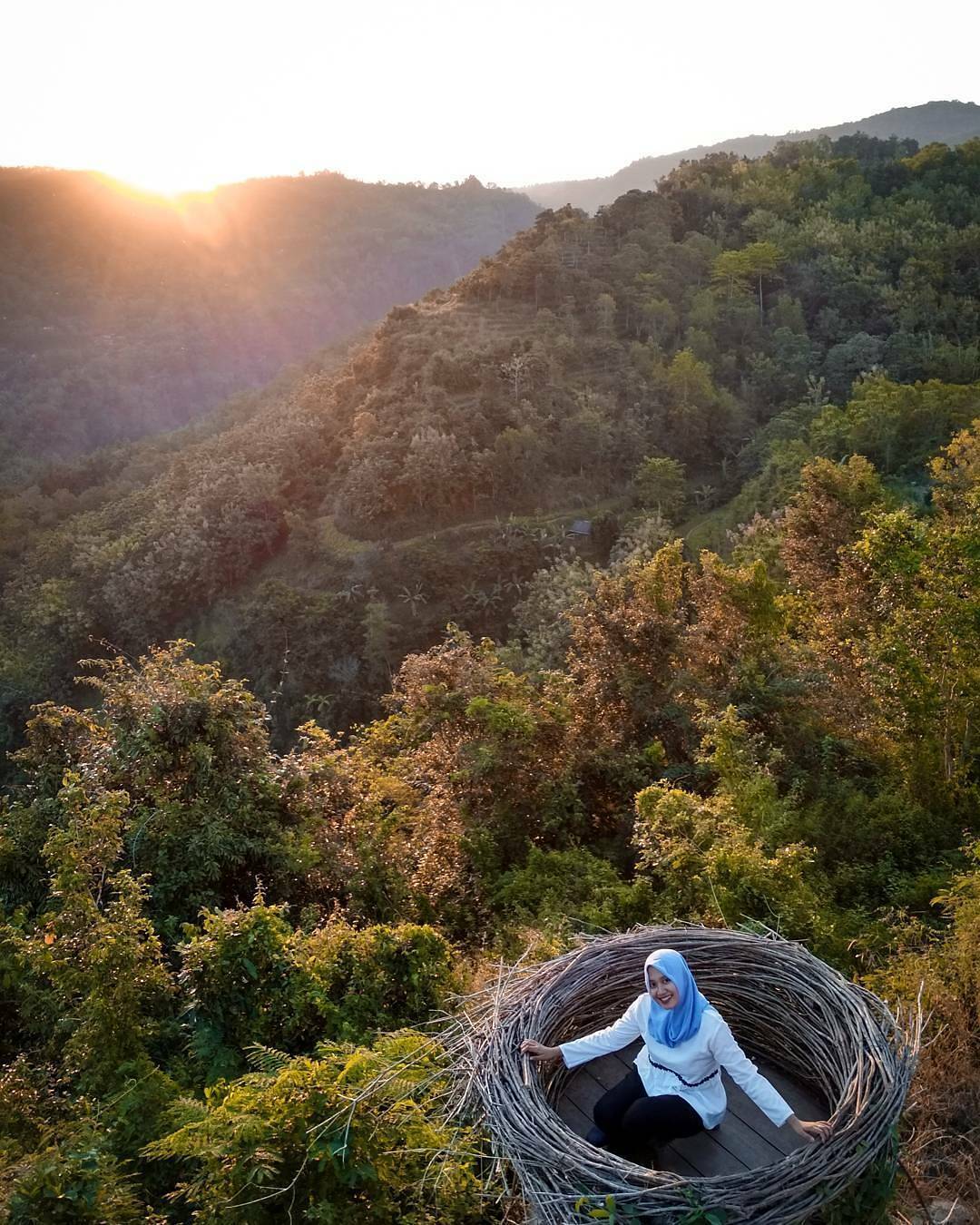 A woman sitting in a giant bird's nest overlooking green hills and a beautiful sunset at Mojo Gumelem Hill in Yogyakarta, Indonesia.