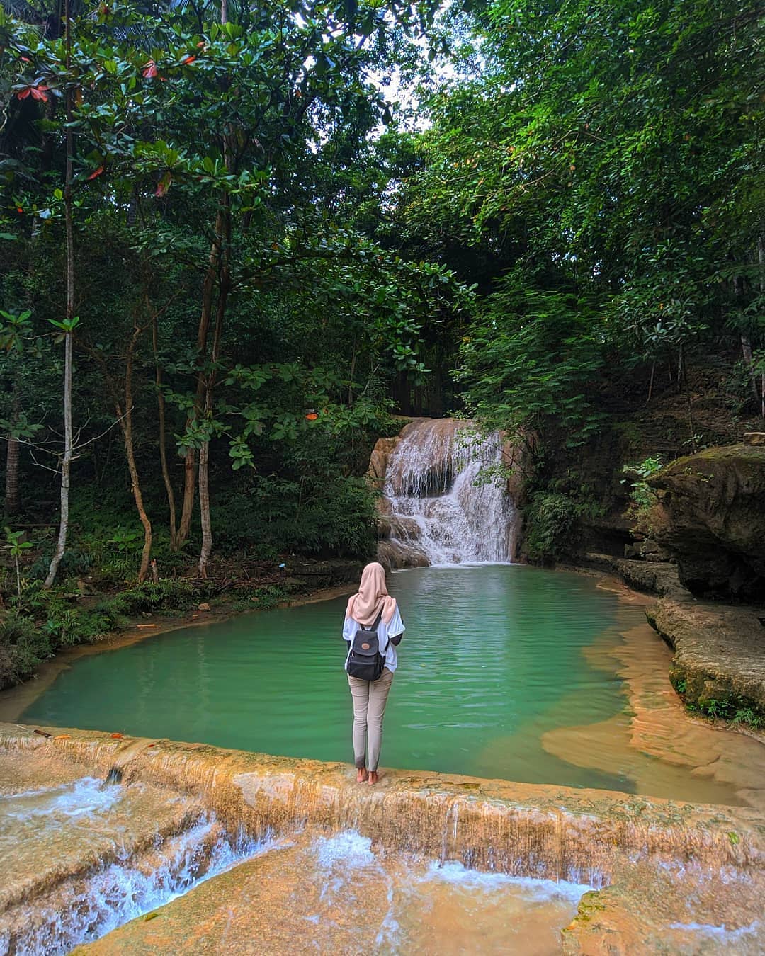 A woman standing by the turquoise pool at Lepo Waterfall, gazing at the cascading waterfall surrounded by lush greenery in Yogyakarta.