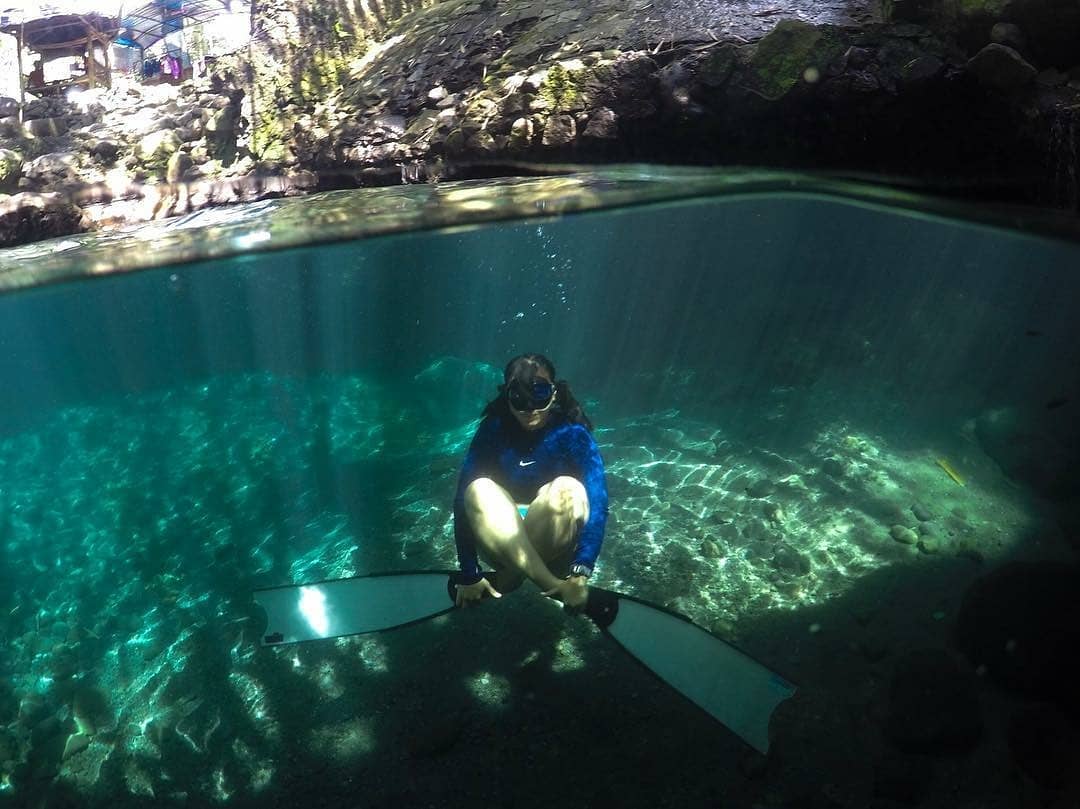 A person wearing snorkeling gear underwater in the clear blue-green waters of Tirta Budi Bathing Place, surrounded by rocky formations and sunlight filtering through the water surface.