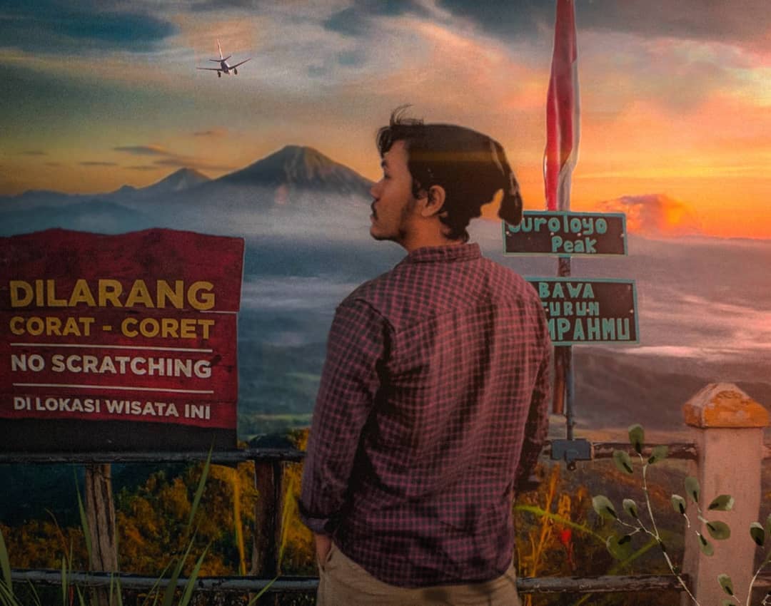 A person standing near signs at Suroloyo Peak in Yogyakarta, Indonesia, looking towards the sunrise over cloudy mountains in the distance. A "No Scratching" sign and a signpost indicating Suroloyo Peak are visible.