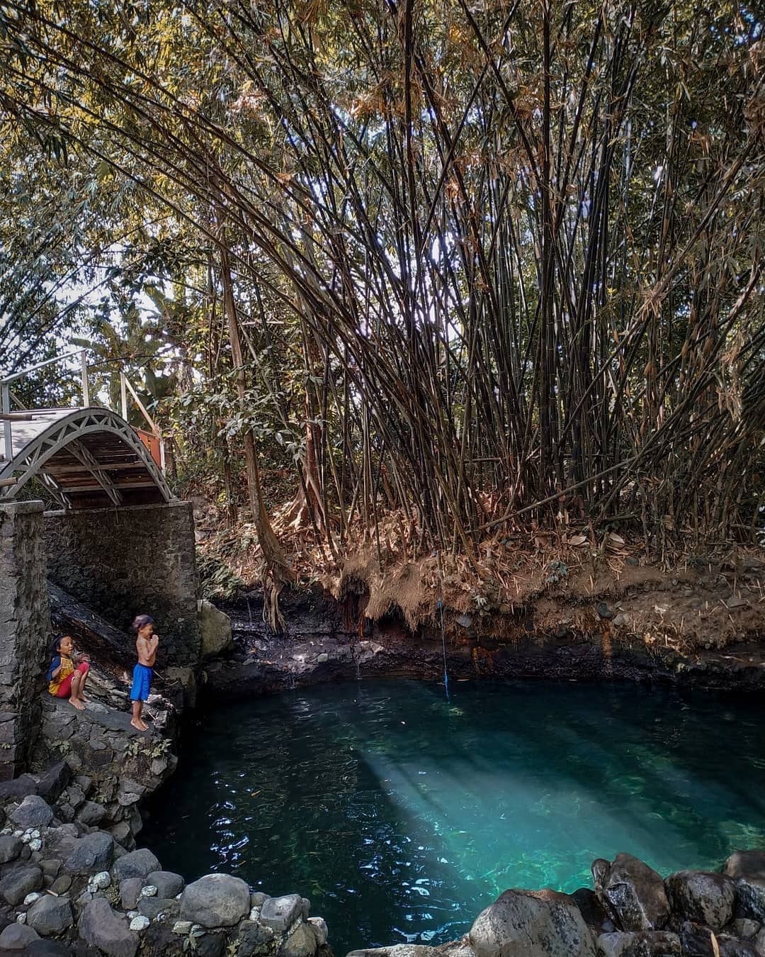 Two children stand at the edge of the clear blue-green waters of Tirta Budi Bathing Place, surrounded by rocks and dense bamboo vegetation with a small bridge in the background.