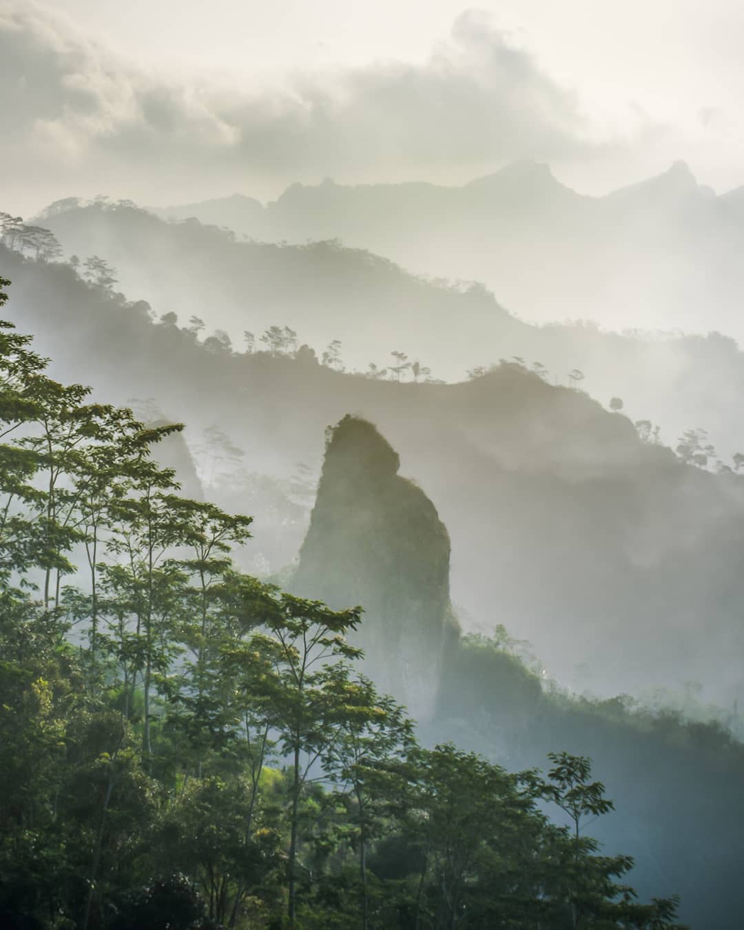 A misty view of the lush green hills surrounding Suroloyo Peak in Yogyakarta, Indonesia, with distinctive rock formations and trees in the foreground.