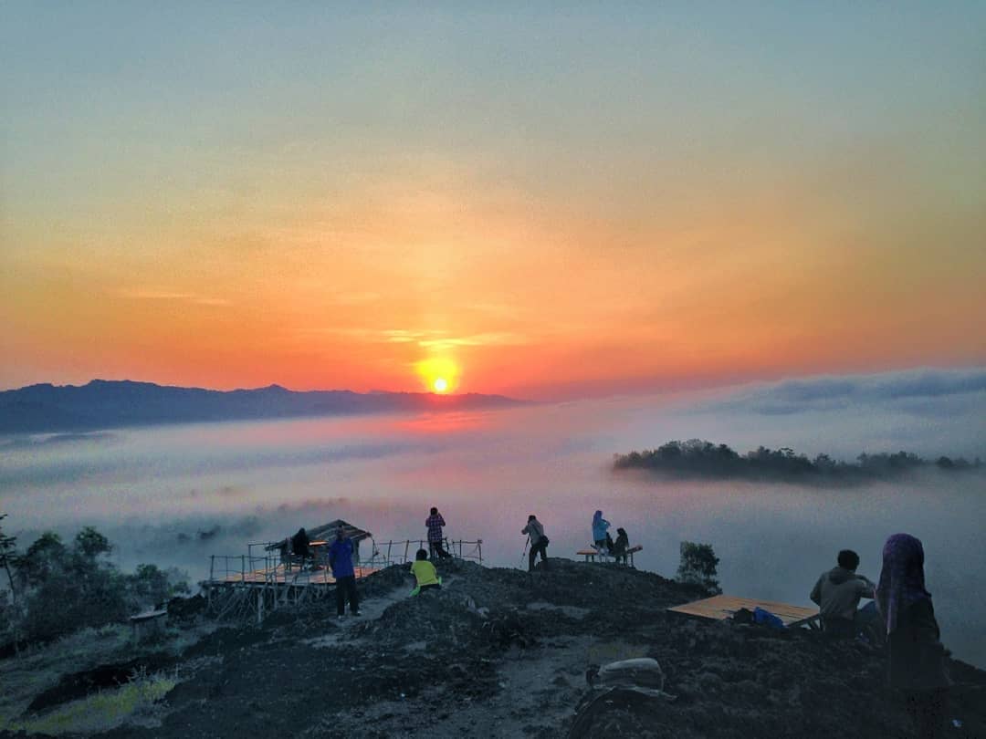 A group of people watching the sunrise from the summit of Ireng Mountain, with a misty landscape spread out below and the sun rising over the horizon.