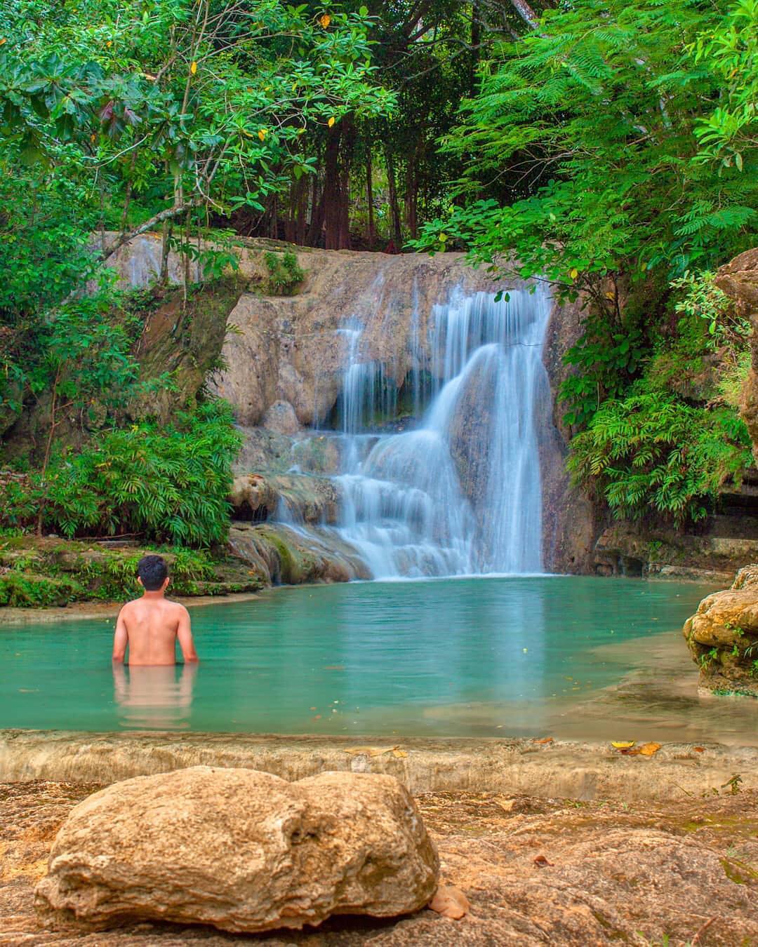 A man standing in the turquoise pool at Lepo Waterfall, surrounded by lush greenery and cascading water in Yogyakarta.