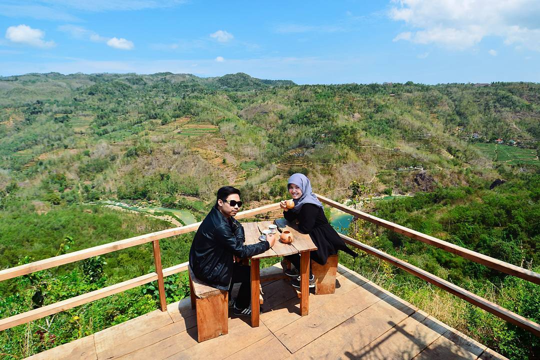 A couple enjoys a cup of coffee at a wooden table overlooking the lush green landscapes from a viewing platform at Mojo Gumelem Hill in Yogyakarta, Indonesia.