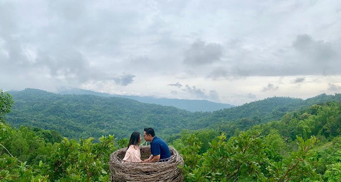 A couple sitting inside a large bird nest-like structure at Green Village Gedangsari with lush green hills in the background.