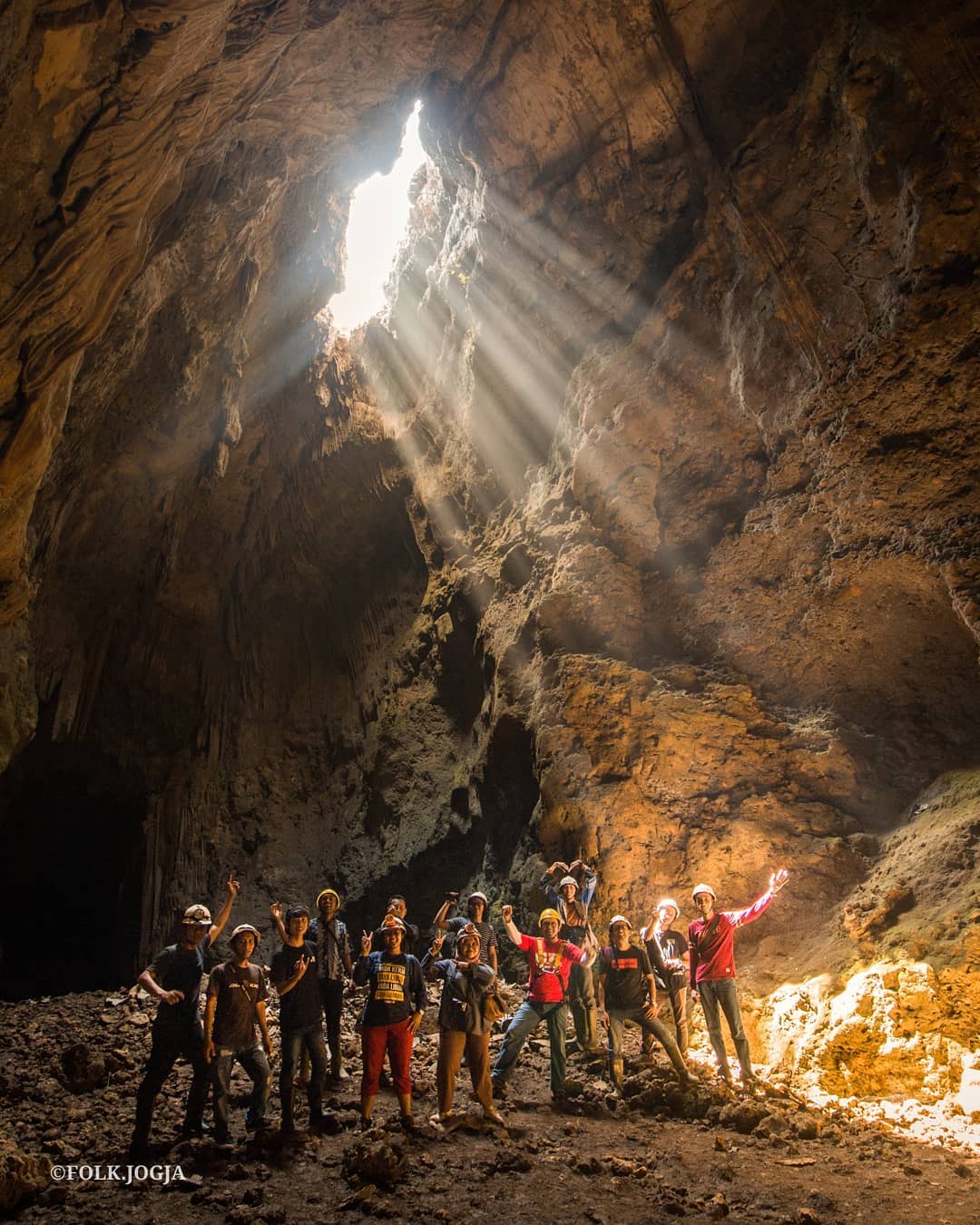 A group of people wearing helmets poses inside Cokro Cave, with sunlight streaming in from above.