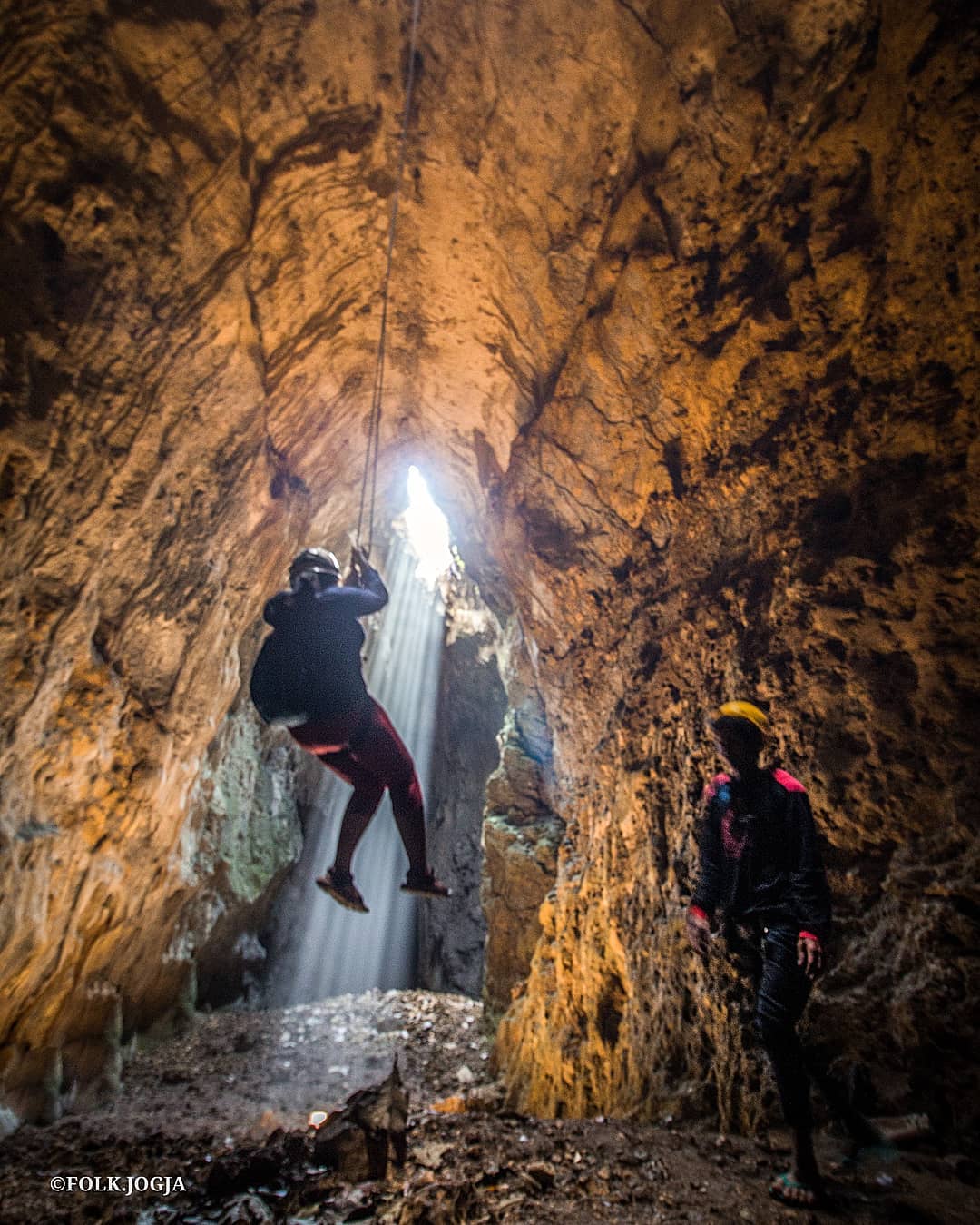 A person rappels down into Cokro Cave while another person watches from below, with sunlight streaming in through the entrance.