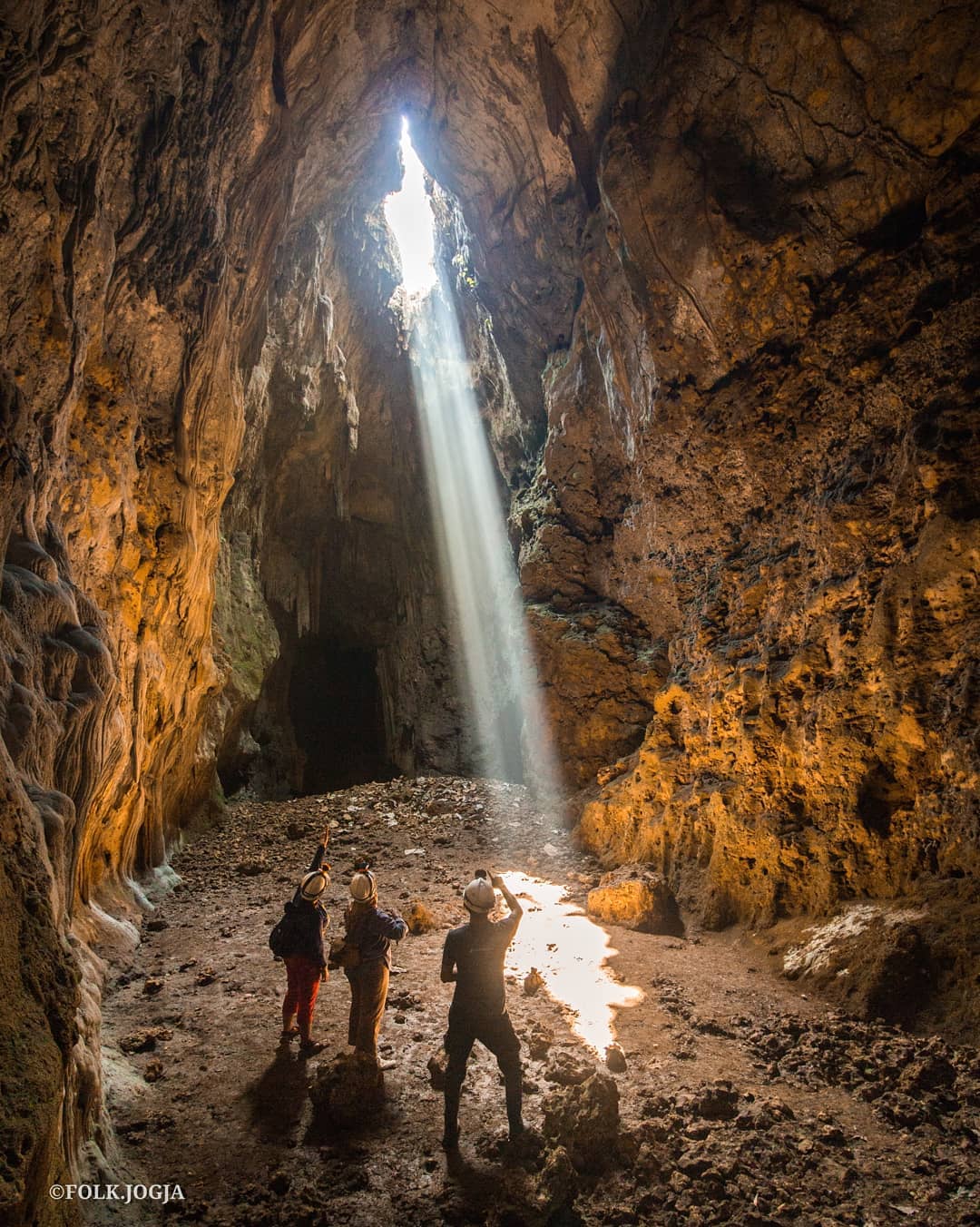 Three people in helmets stand inside Cokro Cave, looking up at a beam of sunlight entering through the cave's vertical shaft.